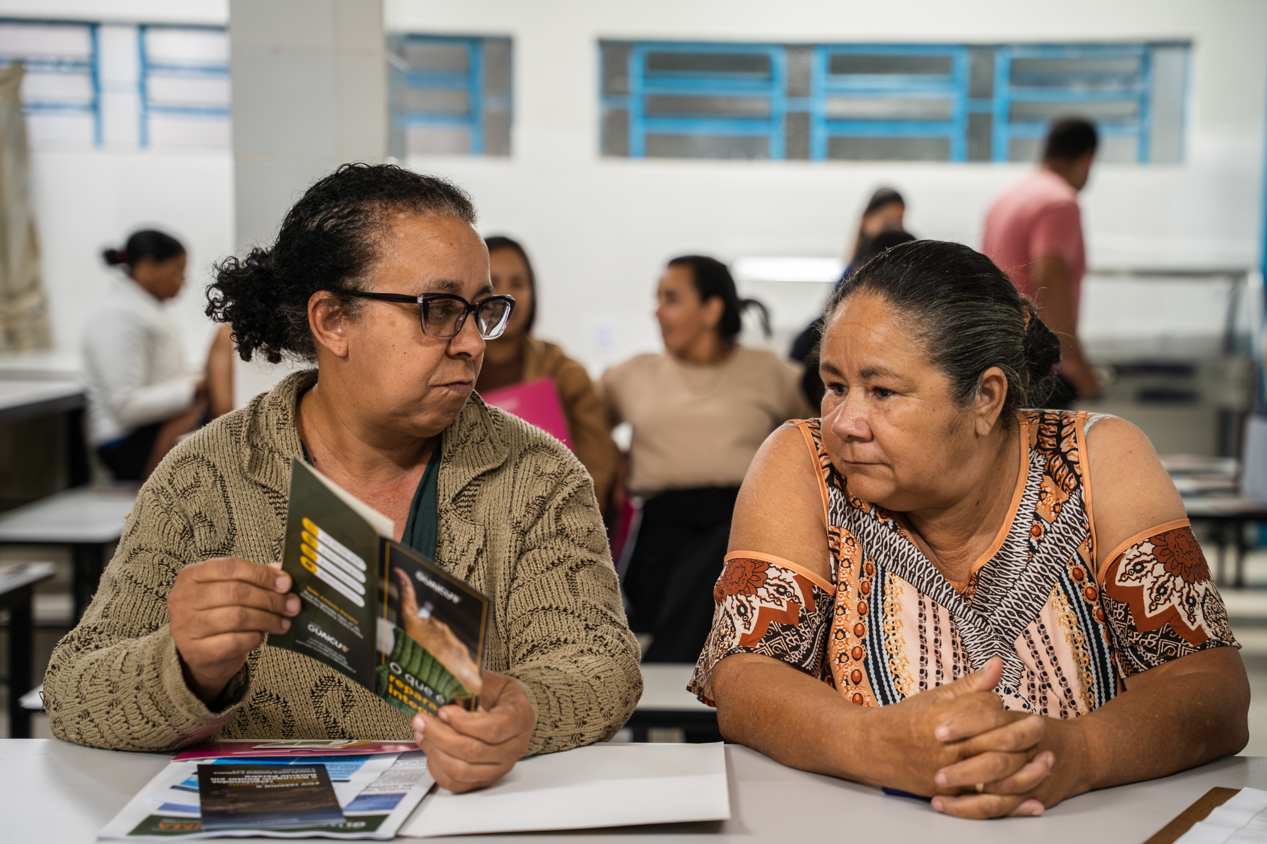 Duas mulheres, atingidas de Antônio Pereira, mostram uma a outra os materiais impressos distribuídos dentro de pastas para os participantes. Ela segura um folder aberto sobre a reparação integral. Dentro da pasta há mais um folder, sobre a FGV e cartilhas sobre o Cadastro. 