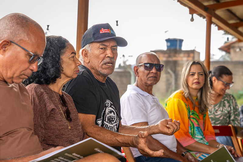 Seis moradoras e moradores de Antônio Pereira sentados em uma roda, discutindo os temas da reunião enquanto seguram pastas com materiais impressos sobre a FGV, reparação e assessoria técnica. 