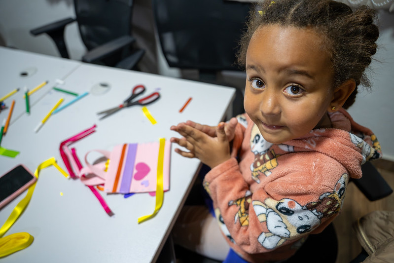 Criança olha diretamente para a câmera enquanto brinca com papéis e fitas coloridas.