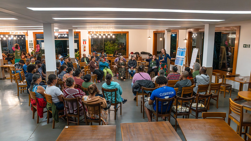 Grupo de moradoras e moradores de Antônio Pereira reunidas em diálogo em uma sala ampla e iluminada. De pé, uma analista do Instituto Guaicuy explica temas relacionados ao Cadastro da FGV com apoio visual de um cavalete com materiais impressos. 