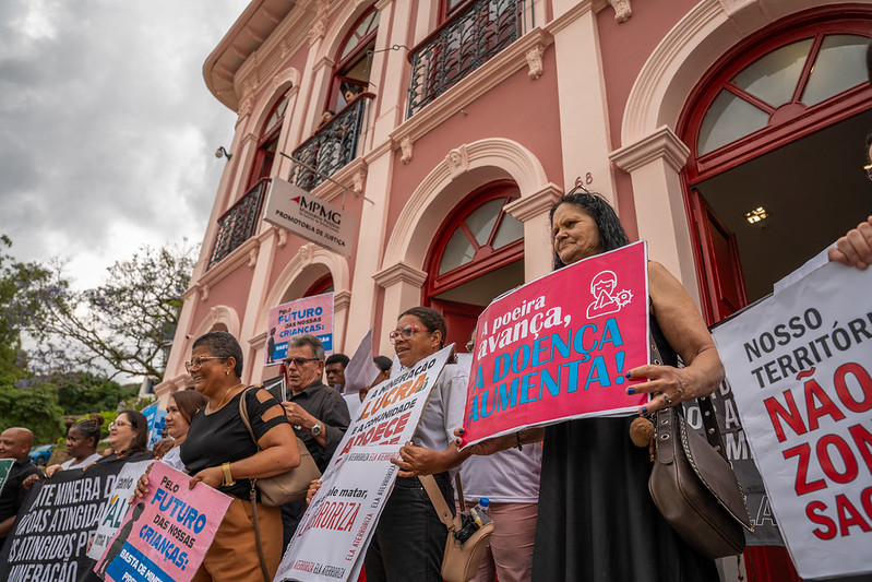 Grupo de pessoas reunidas em frente ao Ministério Público de Minas Gerais em Ouro Preto em manifestação pelos direitos das pessoas atingidas de Antônio Pereira. As pessoas carregam cartazes com os dizeres "nosso território não é zona de sacrifício", "a poeira avança, a doença aumenta", pelo futuro das nossas crianças", dentre outras.