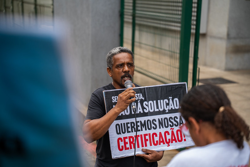 Foto de homem segurando microfone em uma mão e cartaz colorido em outra durante manifestação. O cartaz possui os dizeres "sem resposta, não há solução. Queremos nossa certificação!"