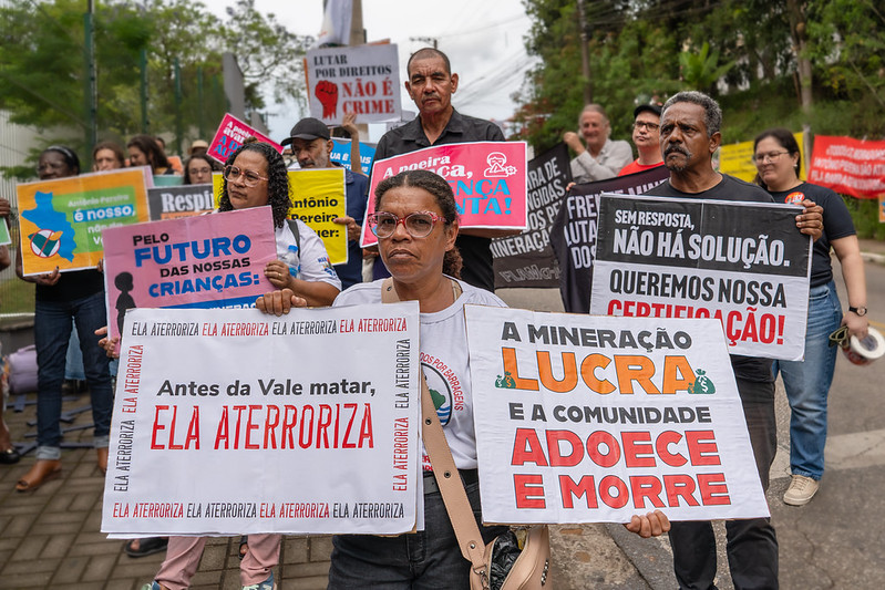Foto de manifestação de pessoas atingidas de Antônio Pereira segurando diversas placas coloridas com os dizeres "Antes da Vale matar, ela aterroriza", "a mineração lucra e a comunidade adoece e morre", "pelo futuro das nossas crianças", 'sem resposta não há solução, queremos nossa certificação", dentre outras.