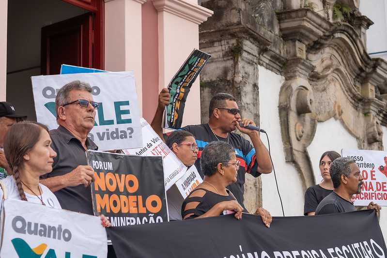 Foto mostra homem falando em microfone entre outras pessoas carregando faixas e cartazes em manifestação de pessoas atingidas de Antônio Pereira, realizada em frente ao Ministério Público de Minas Gerais, em Ouro Preto. As placas carregam dizeres como "Por um novo modelo de mineração", "Quanto Vale uma vida?", dentre outras.