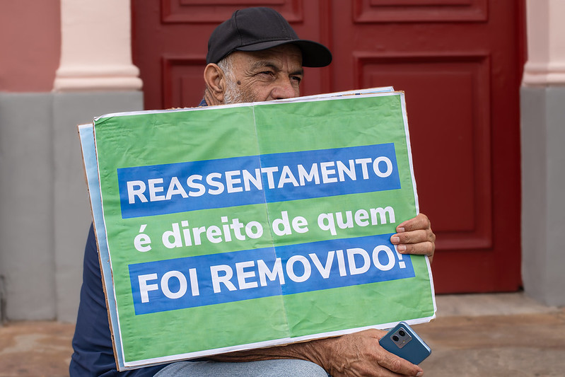 Foto de homem segurando cartaz verde com os dizeres "reassentamento é direito de quem foi removido!" durante manifestação de pessoas atingidas de Antônio Pereira.