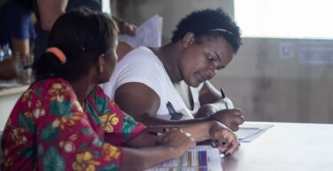 foto de duas mulheres quilombolas em reunião sobre o Acordo de Reparação das Pessoas Atingidas pela Vale. Elas estão sentadas, a primeira delas segura um informativo e a segunda escreve em um caderno