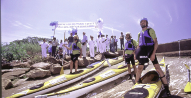 Participantes da Expedição Manuelzão utilizando equipamento de segurança posam ao lado dos caiaques, no rio, enquanto às margens um grupo de pessoas com roupas brancas segura balões e uma faixa que pela preservação do Rio das Velhas.