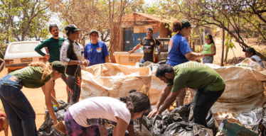 Grupo de pessoas com adultos e crianças, reunido em um espaço ao ar livre em espaço rural, manuseando grandes sacos pretos com os materiais recolhidos. Ao fundo, há árvores, carros e uma pequena construção simples.