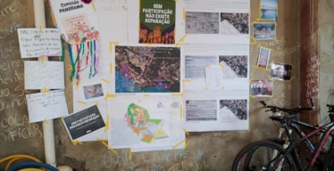 Um mural exibe mapas, fotos, cartazes e anotações relativos ao mapeamento social participativo. Na parede ao redor, há textos escritos à giz reinvindicando direitos relativos à reparação e duas bicicletas encostadas.