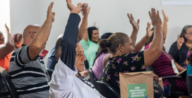 Grupo de pessoas sentadas em cadeiras, em uma sala, levantam as mãos, aparentemente em uma votação ou expressão de apoio em uma reunião. Em primeiro plano, um homem com uma camiseta branca também segura um saco de papel com uma imagem impressa colada escrito "Sem participação não existe reparação"