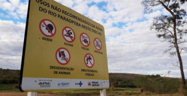 Placa amarela área externa aberta onde está escrito "Usos não recomendados da água do Rio Paraopeba neste trecho". Abaixo do texto, ícones ilustram as proibições, que incluem: beber, nadar, pescar, irrigação, dessedentação de animais e abastecimento público. Há também os logotipos de instituições responsáveis, como MPMG, Igam, SUS e Estado de Minas Gerais.