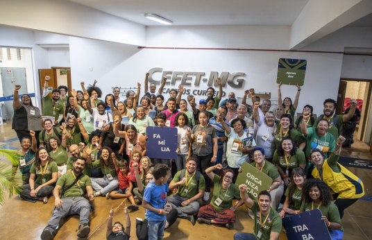 Grupo grande e diverso de pessoas, com algumas delas uniformizadas, posando para a foto. Muitas estão sorrindo e levantando os braços em gesto de celebração. Algumas seguram placas com palavras como "família", "organização" e "trabalho".