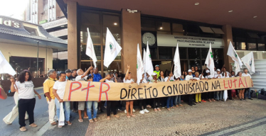 Grupo de pessoas reunido em frente ao edifício do Ministério Público de Minas Gerais. Pessoas seguram bandeiras e uma grande faixa escrito: "PTR Direito conquistado na luta!". O ambiente é iluminado pela luz do dia e o grupo parece participar de uma manifestação.