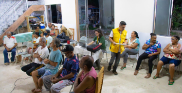 Grupo de pessoas em reunião comunitária em espaço interno, com participantes sentados e interagindo.