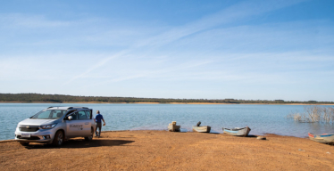 Paisagem diurna em área aberta com um rio, quatro barcos simples próximos à margem, um carro com adesivo escrito Guaicuy e um homem de costas.