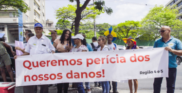 Grupo de pessoas participando de uma manifestação ao ar livre, em um local urbano, durante o dia. Parte das pessoas seguram uma faixa onde se lê: "Queremos perícia dos nossos danos! Região 4 e 5".