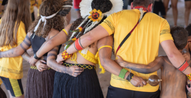 Várias pessoas, fotografadas de costas, se abraçam em roda. A maioria usa blusas amarelas e acessórios indígenas.