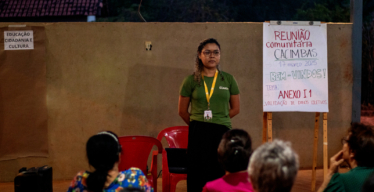 Mulher em pé uniformizada ao lado de flipchat fala para pessoas sentadas em cadeiras que aparecem de costas