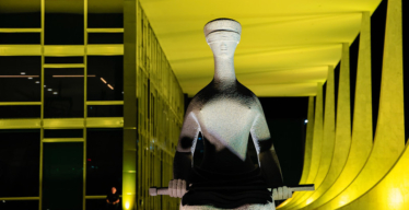 Fachada do prédio do Supremo Tribunal Federal (STF) à noite, iluminada de amarelo, com a escultura "A Justiça" em primeiro plano.