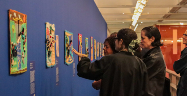 Um grupo de pessoas, especialmente mulheres, observa bordados expostos em parede.