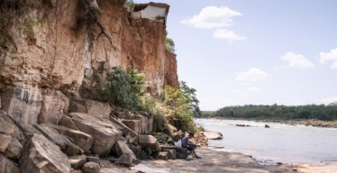 Imagem mostra um barranco alto à beira do rio com um imóvel degradado no topo, em risco de desabamento. Abaixo, uma criança está sentada sozinha sobre pedras.