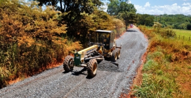 A imagem mostra uma motoniveladora em uma estrada não pavimentada.