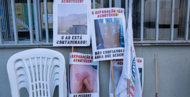 Cadeiras vazias empilhadas ao lado de cartazes colados em suportes de madeira com os seguintes escritos: "A reparação não aconteceu! O ar está contaminado!", "A reparação não aconteceu! Não congiamos na água que bebemos!", "A reparação não aconteceu! As pessoas atingidas estão doentes!".