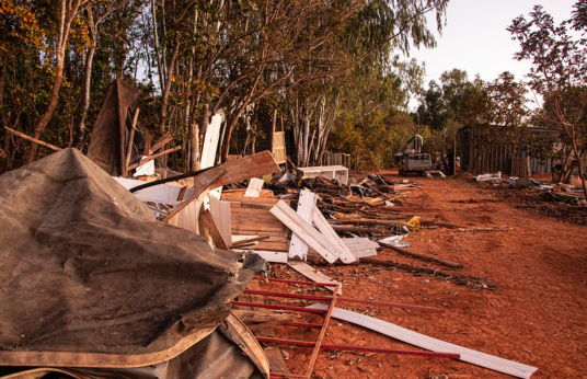 Destroços de construção em chão de terra, com veículo para demolição ao fundo.