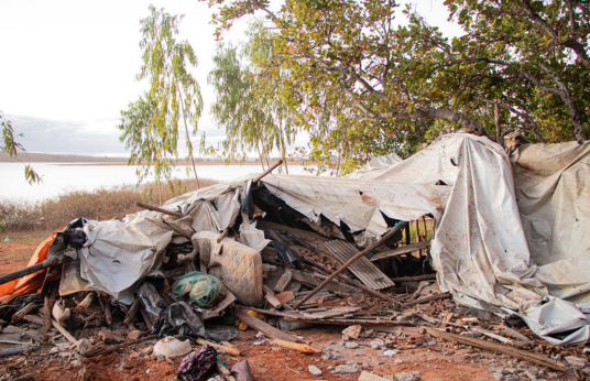 Destroços de construção e itens pessoais danificados próximos a vegetação e represa