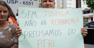 Em espaço aberto, em frente a grupo de pessoas, mulher segura cartaz escrito "Chega de promessas. Lei é só para a Vale? PTR auxílio obrigatório".