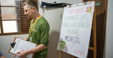 Homem jovem em local fechado, voltado para outra pessoa fora do quadro, próximo a flipchart com o texto "Bem vindas à formação das funções da Instância Regional — Mediação".