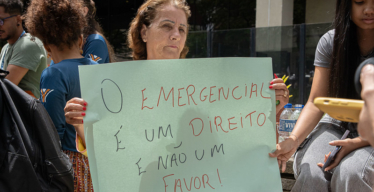 Mulher idosa em espaço aberto, próximo a outras pessoas, segura cartaz escrito "o Emergencial é um direito e não um favor!"