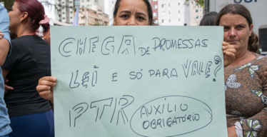 Em espaço aberto, em frente a grupo de pessoas, mulher segura cartaz escrito "Chega de promessas. Lei é só para a Vale? PTR auxílio obrigatório".