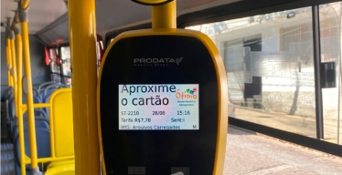 Interior de ônibus com mão encostando o cartão em sistema de bilhetagem eletrônica. O valor da passagem é R$7,70.
