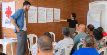 Plateia sentada em cadeiras assiste a apresentação de uma dupla formada por um homem e uma mulher com base em cartazes colados em uma parede.