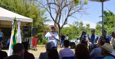 Homem idoso utilizando um chapéu fala, de pé, para público sentado. O espaço é próximo à natureza e aberto, mas há tendas. Também há bandeiras, indicando formalidade.