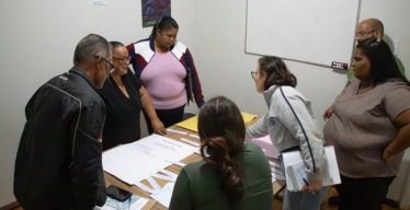 Grupo de homens e mulheres olha materiais em mesas em sala fechada, em frente a um quadro branco.