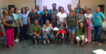 Grupo de pessoas em pé, algumas uniformizadas, em ambiente fechado, em frente a telão.
