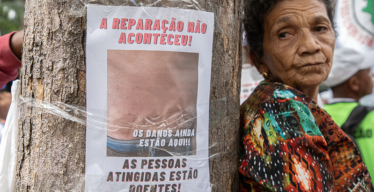 Cartaz fixado em árvore diz "a reparação não aconteceu. As pessoas atingidas estão doentes" e mostra uma foto de pele com reação alérgica. Ao lado, mulher idosa parece cansada.