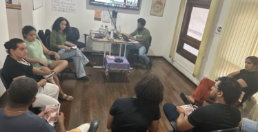 Pessoas em ambiente fechado, algumas uniformizadas, sentadas em roda. Um das pessoas usa um computador e tela grande mostra uma apresentação.