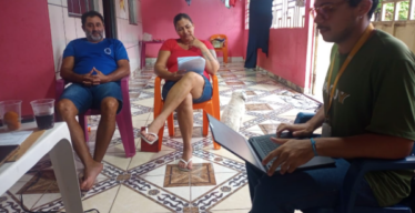 Três pessoas sentadas na varanda de uma casa. Uma delas, com o uniforme do Instituto Guaicuy, utilizando um computador.