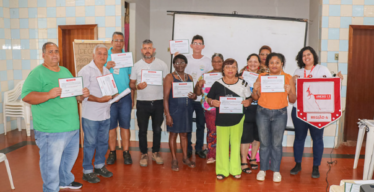 Pessoas em ambiente interno posam para foto com certificados em mãos. Uma delas segura um estandarte escrito "Anexo 1.1 Região 4".