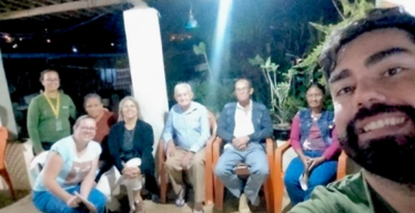 Várias pessoas posam para foto em espaço aberto coberto durante a noite. Ao fundo, uma delas, de pé, utiliza uniforme e crachá. Em primeiro plano, aparece o rosto de outra pessoa de uniforme, que está tirando a foto.