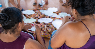 Grupo de mulheres sentadas em roda. Duas delas, em primeiro plano, usam blusas roxas e fazem crochê.
