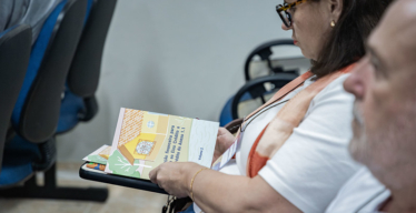 Mulher de perfil sentada em cadeira em ambiente fechado manuseia uma cartilha impressa