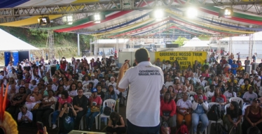 Foto de Guilherme Boulos em palco, de costas, falando ao microfone para a público. Na blusa dele está escrito "Governo do Brasil na rua".