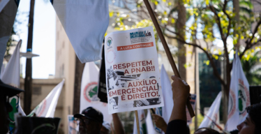Grupo de pessoas em ambiente aberto durante o dia. Algumas empunham bandeiras. Uma delas levanta um cartaz escrito: "Respeitem a PNAB. Auxílio emergencial é direito", no centro da imagem.