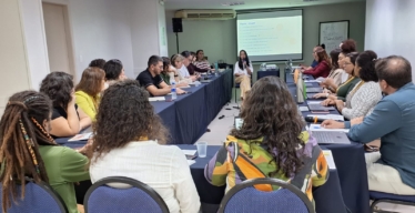 Várias pessoas sentadas em uma mesa de reunião assistindo a uma mulher no centro, que apresenta.