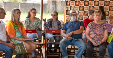 Duas mulheres e um homem sentados em cadeiras de madeira enquanto conversam em grupo.